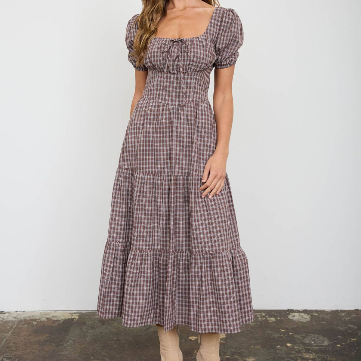 A woman modeling a brown and beige gingham midi dress with puff sleeves, a square neckline, and a tiered skirt, paired with beige ankle boots, set against a plain white wall and textured concrete floor.