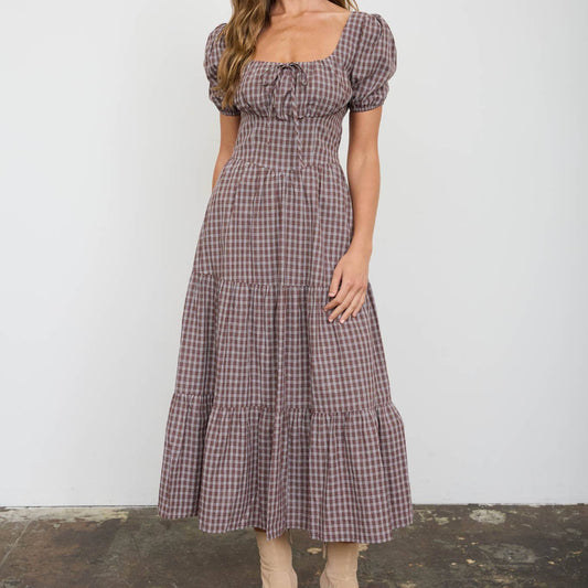 A woman modeling a brown and beige gingham midi dress with puff sleeves, a square neckline, and a tiered skirt, paired with beige ankle boots, set against a plain white wall and textured concrete floor.