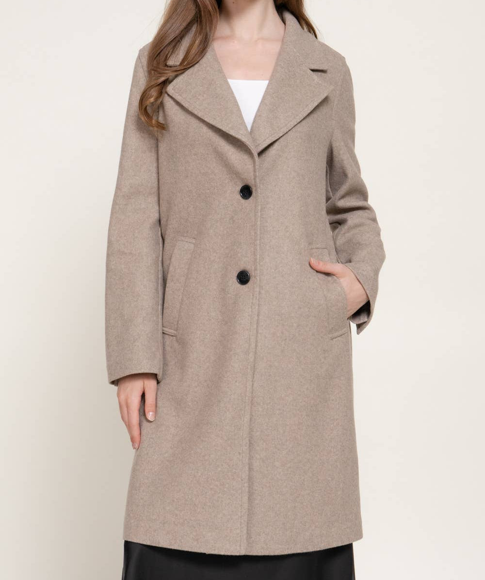 A woman modeling a mid-length, double-breasted overcoat in a warm taupe-beige color, styled over a white top and black skirt against a plain light background.