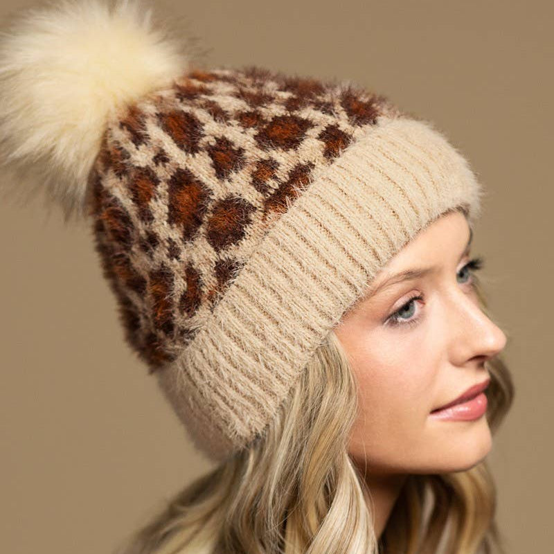 A woman in profile wearing a fuzzy knit beanie with a leopard print pattern and a large cream pom-pom, against a solid beige background.
