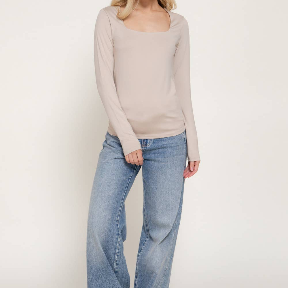 A woman modeling a light beige long-sleeve knit top with a square neckline, paired with light-wash wide-leg jeans, against a plain off-white studio background.