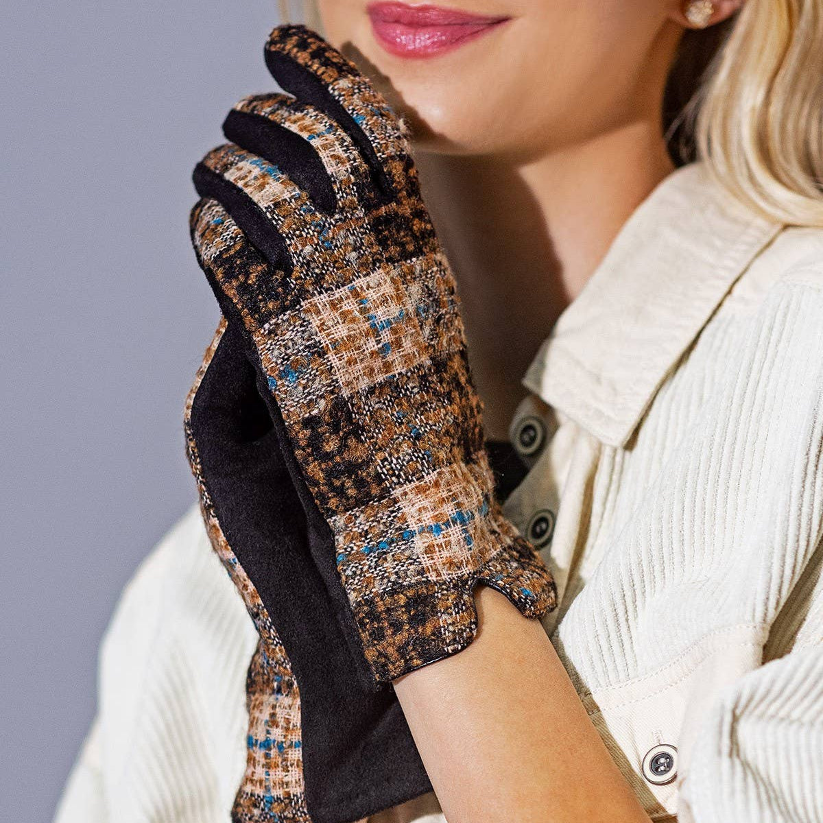 A blonde woman wearing a cream-colored corduroy jacket and holding up a pair of plaid tweed touchscreen gloves with black palms, posing against a solid gray studio background.