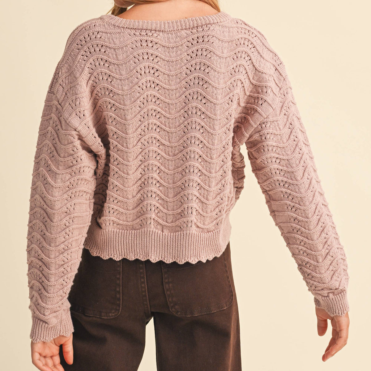 A woman shown from the back wearing a dusty pink textured knit sweater with a wavy chevron pattern and dark brown pants, set against a solid cream background.