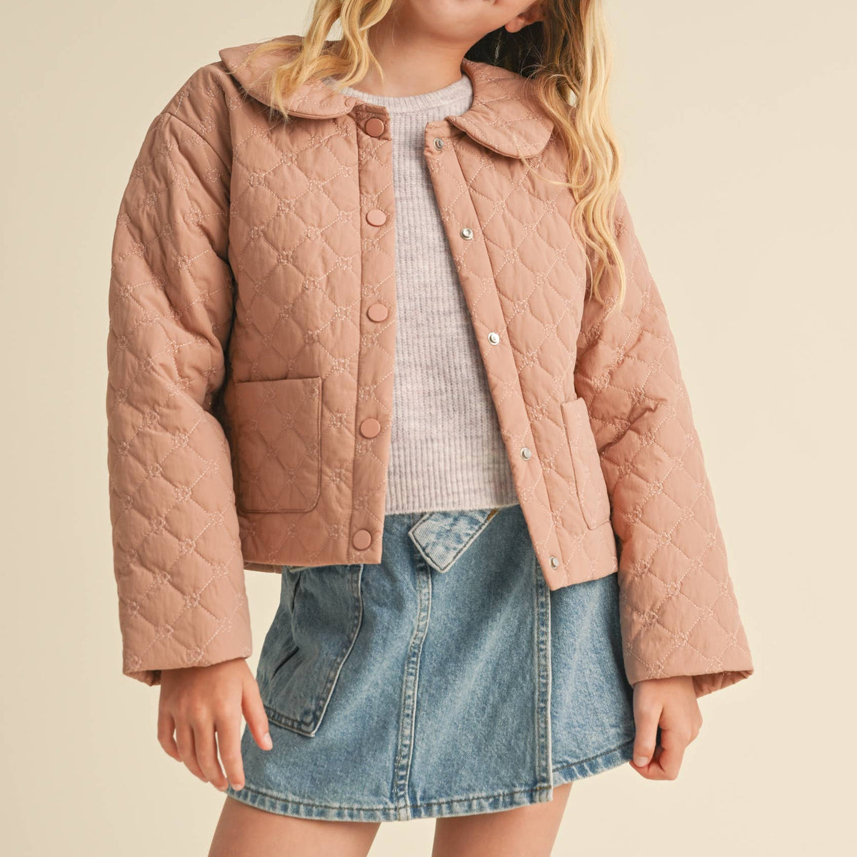 A young girl modeling a quilted cropped jacket in a dusty rose pink color, styled with a light gray knit top and a denim mini skirt, against a solid beige background.
