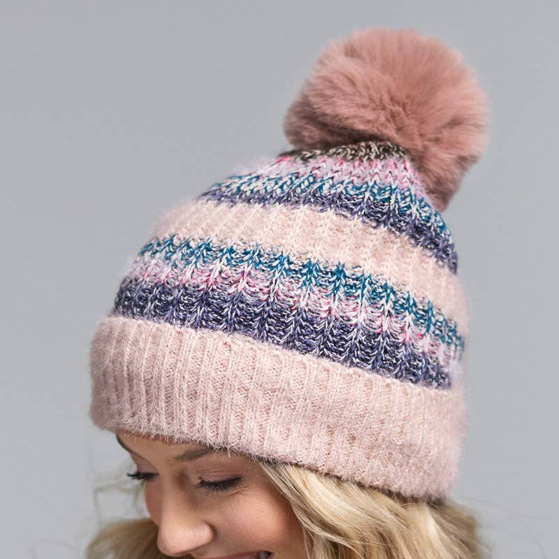 A woman wearing a pink and blue striped knit beanie with a fluffy pom-pom on top, photographed against a plain gray studio background.