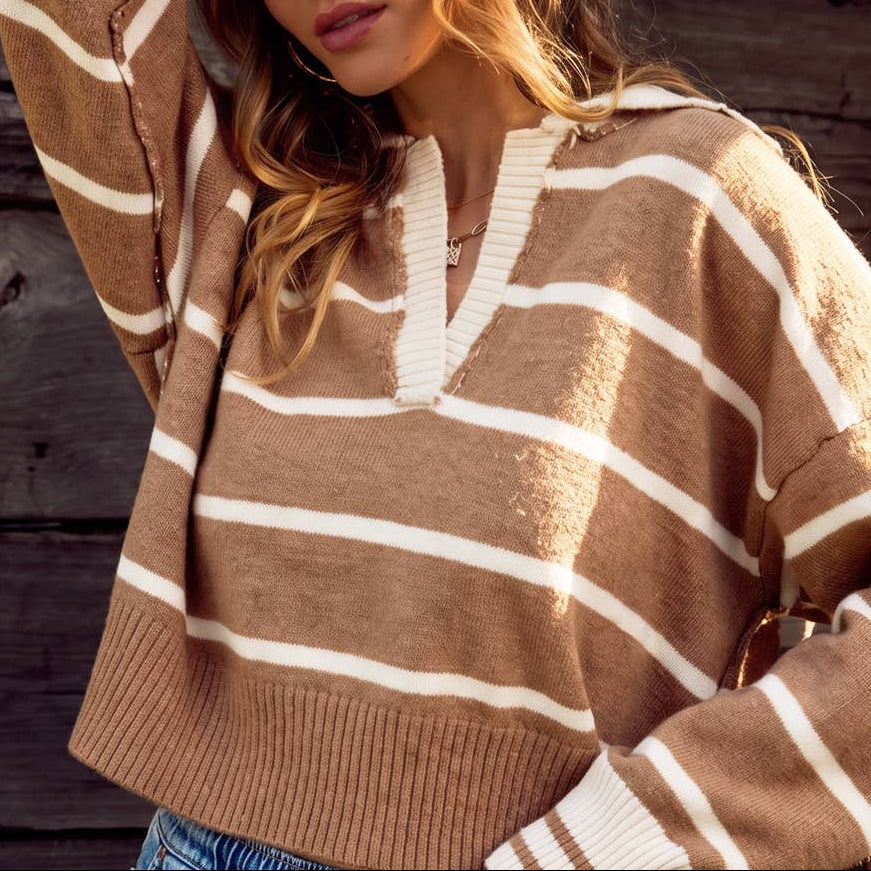 A woman wearing a brown and cream striped knit sweater with a split V-neck, posing against a rustic dark wood plank wall.