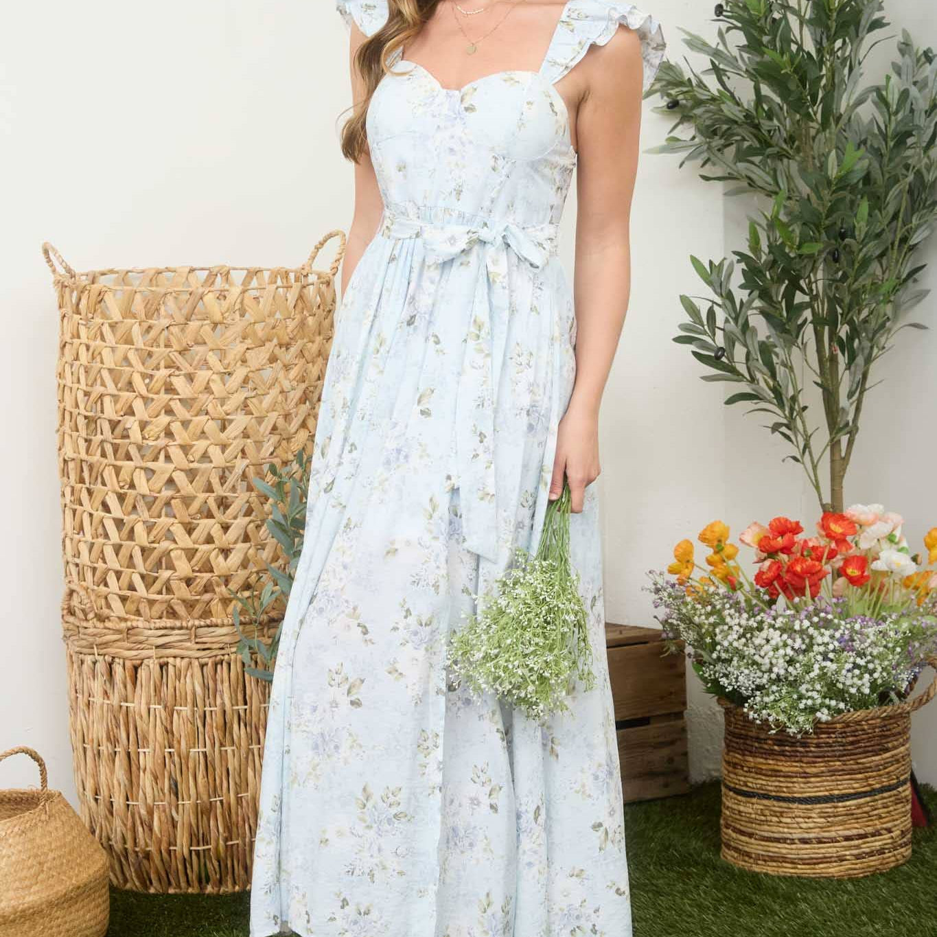 Woman in a light blue floral dress standing outdoors with plants and a woven basket in the background.
