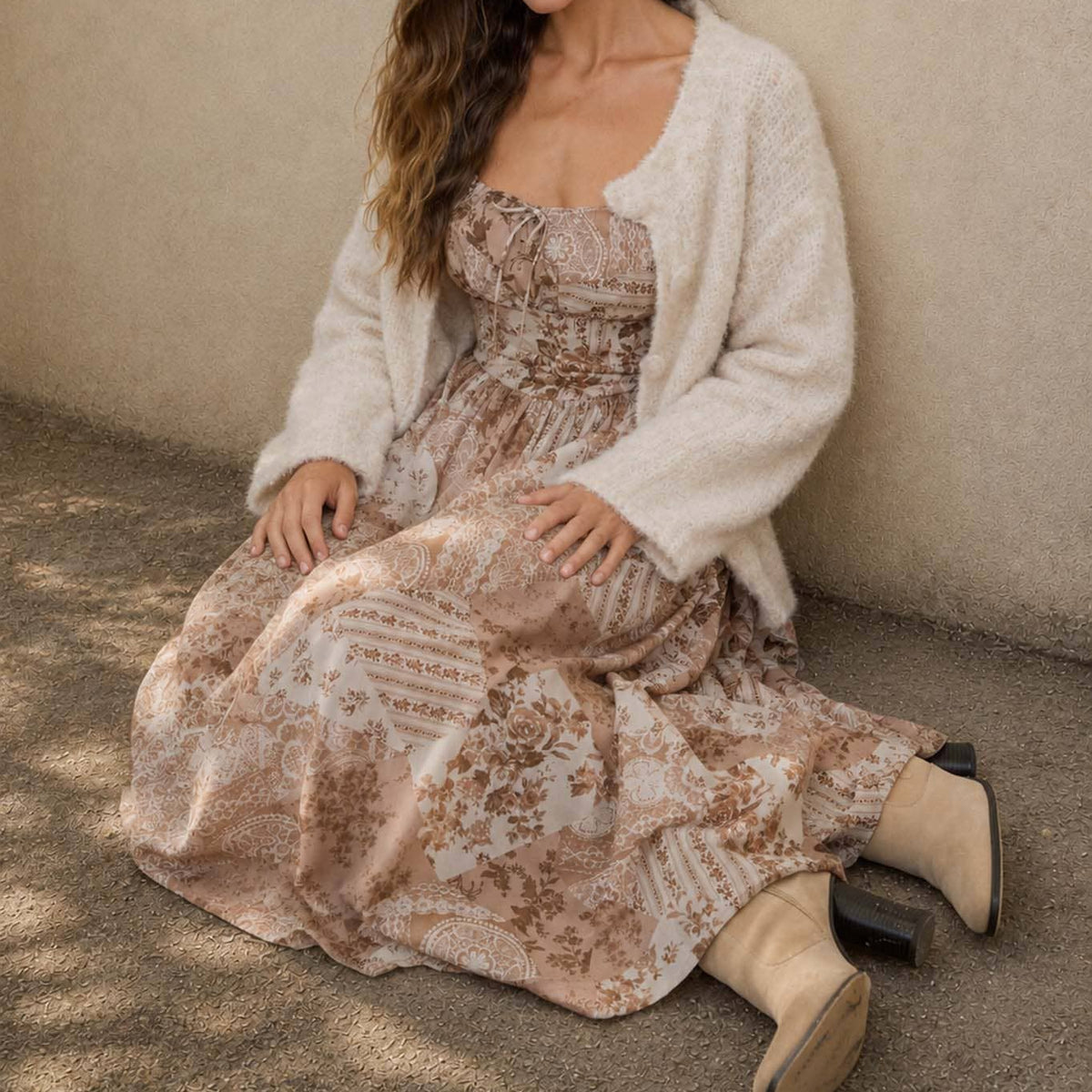 A woman sits on a concrete floor against a textured beige wall, wearing a long-sleeve cream knit cardigan over a strapless midi dress with a brown and beige botanical pattern, paired with beige ankle boots.