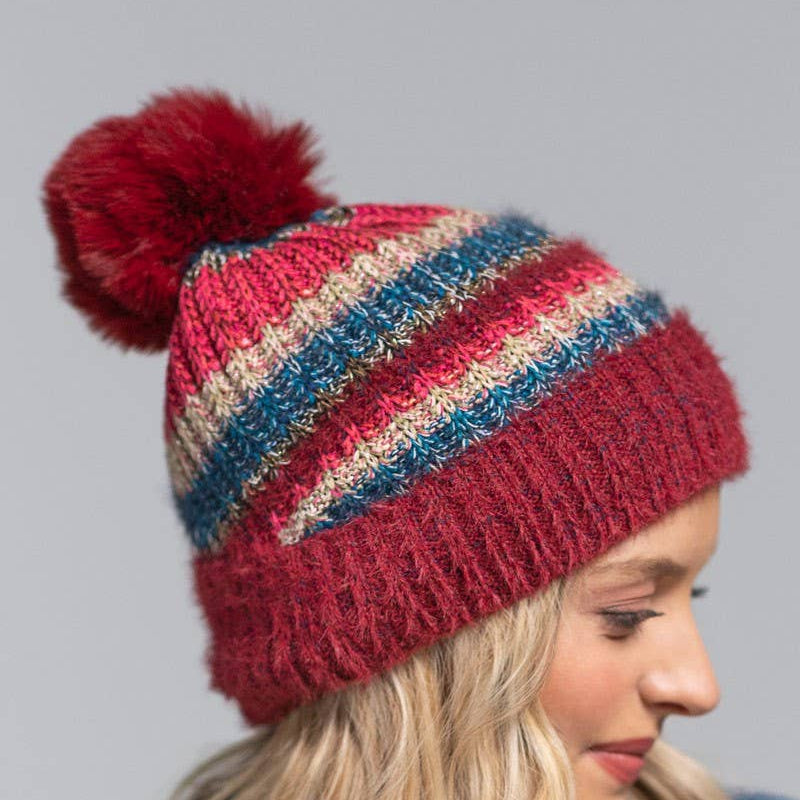 A woman wearing a chunky knit pom-pom beanie with horizontal stripes in burgundy, red, blue, and beige, set against a solid gray background.