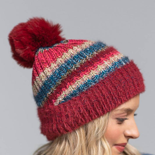 A woman wearing a chunky knit pom-pom beanie with horizontal stripes in burgundy, red, blue, and beige, set against a solid gray background.