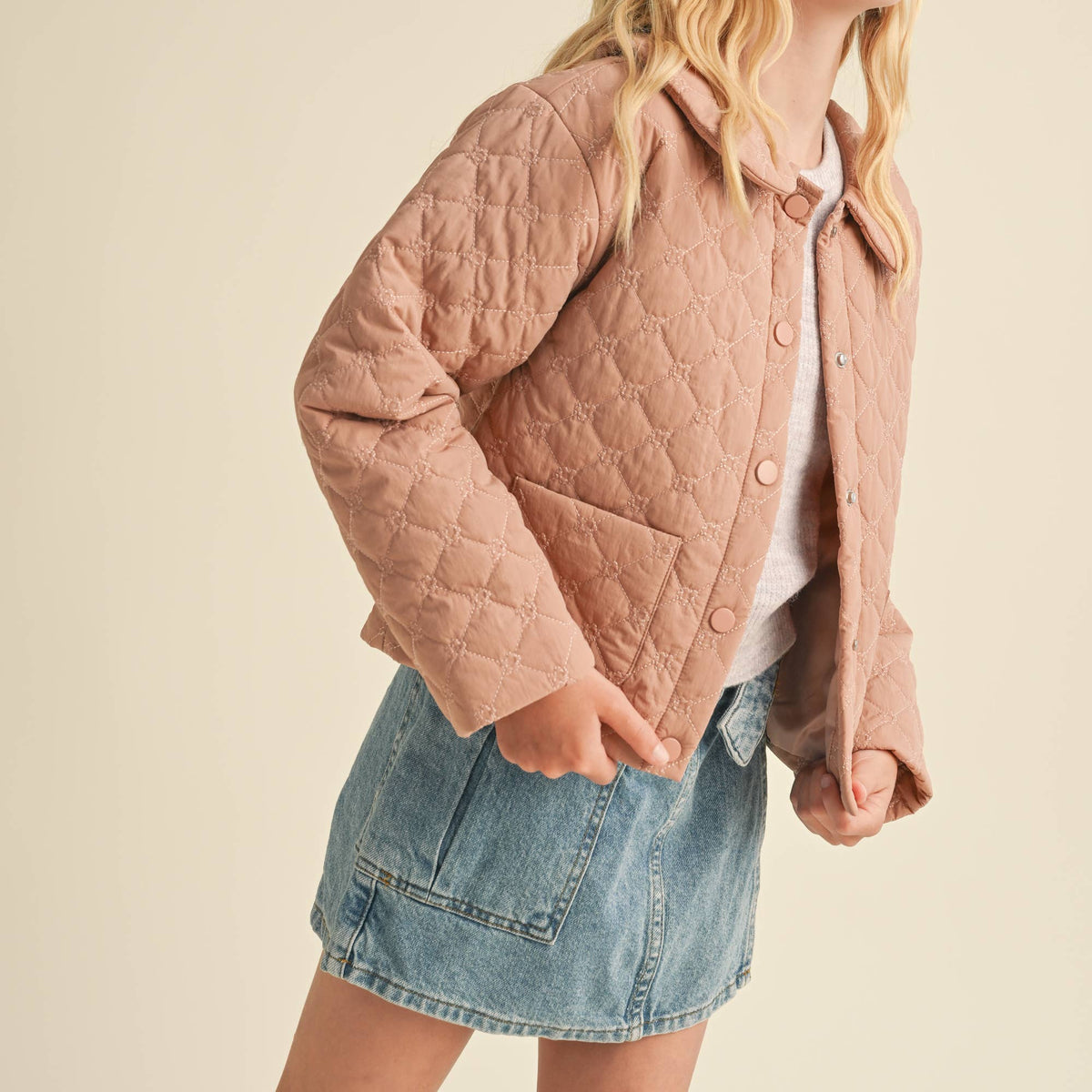 A young girl with blonde hair wearing a dusty pink quilted jacket, a white t-shirt, and a denim mini skirt, posing against a solid beige background.