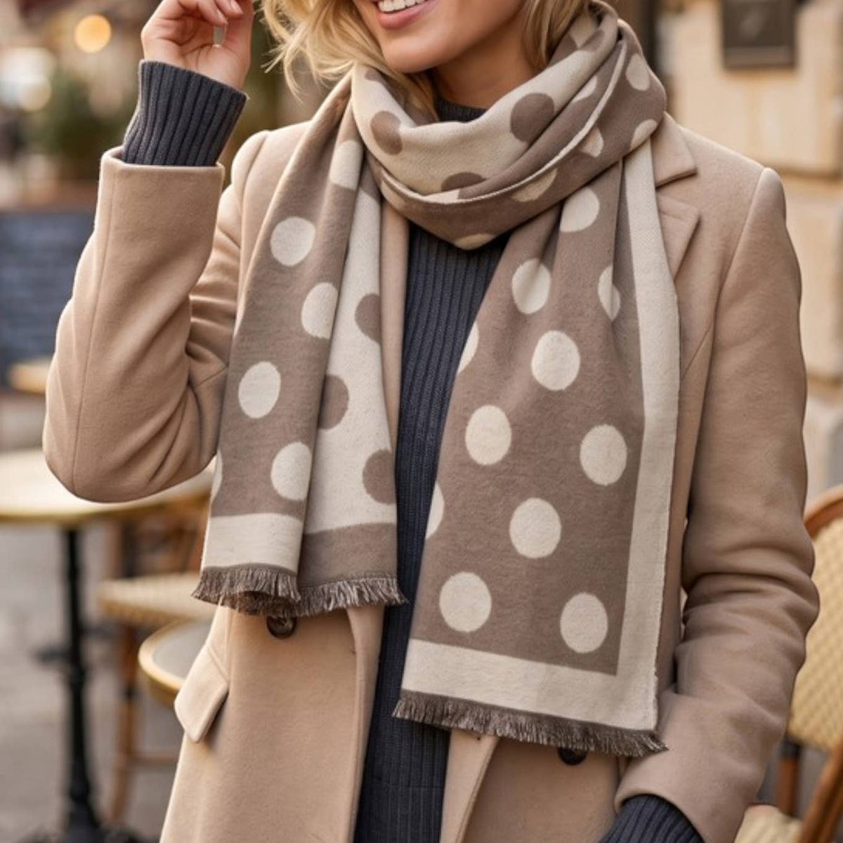 A woman wearing a beige coat and a beige scarf with large white polka dots, standing outdoors on a city street with cafe tables in the background.