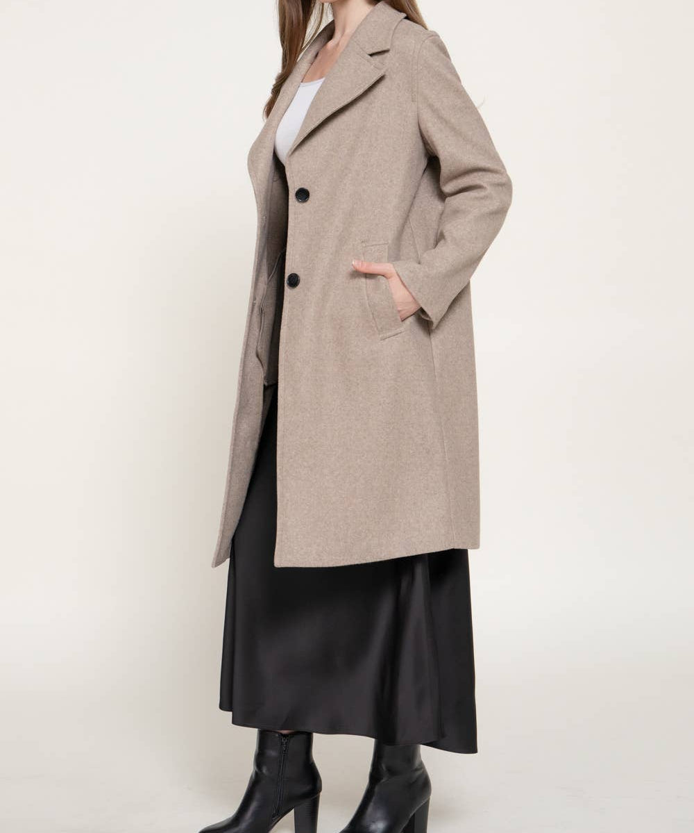 A woman modeling a long, double-breasted beige coat over a white top, black skirt, and black ankle boots, photographed against a plain light background.