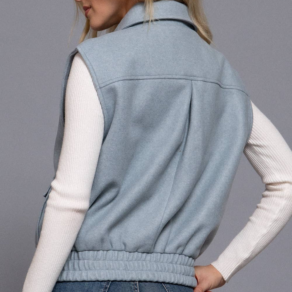 Woman wearing a light blue vest over a white long-sleeve shirt and jeans on a gray background