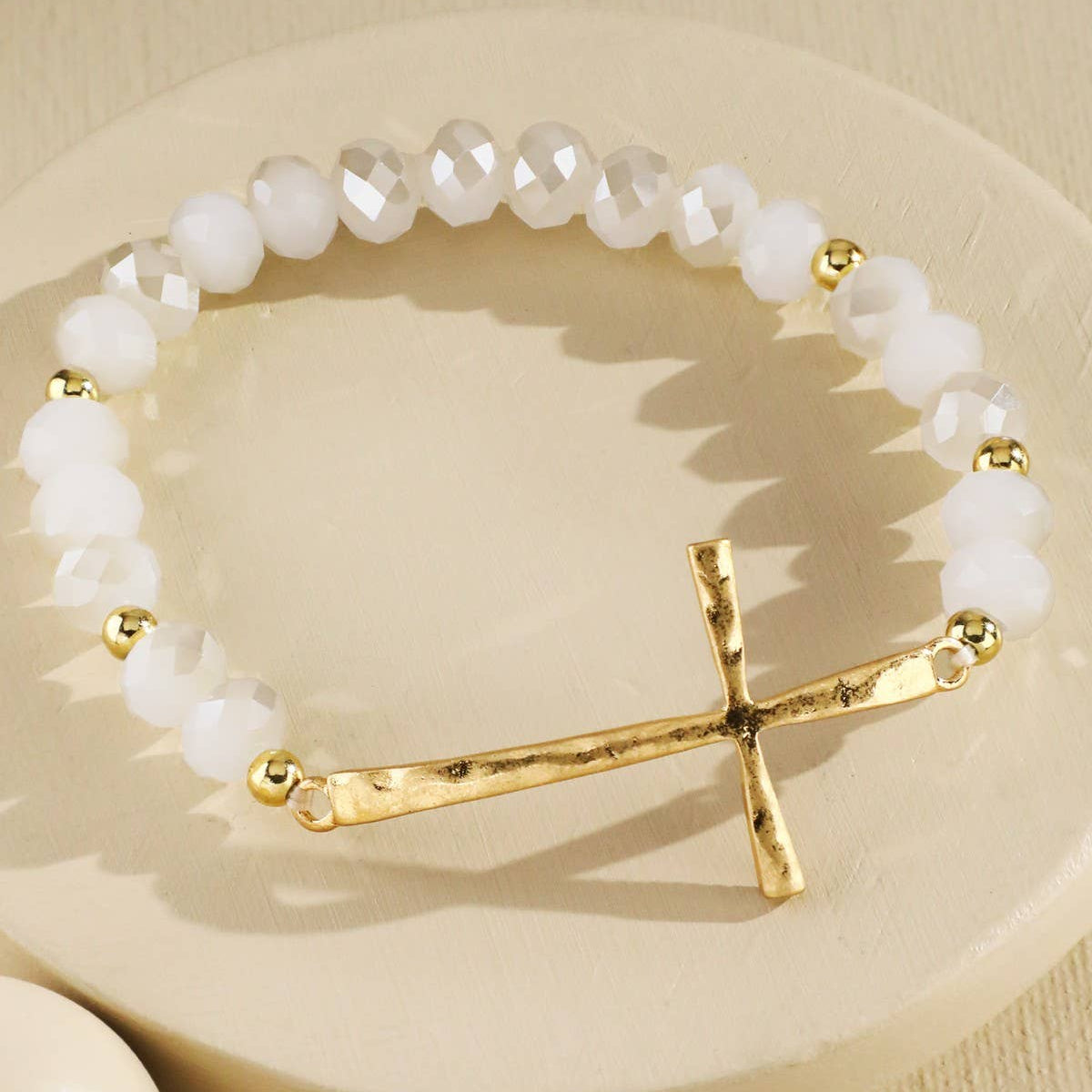 A beaded stretch bracelet featuring white matte beads, faceted white crystal beads, small gold spacer beads, and a hammered gold cross charm, displayed on a round beige pedestal.