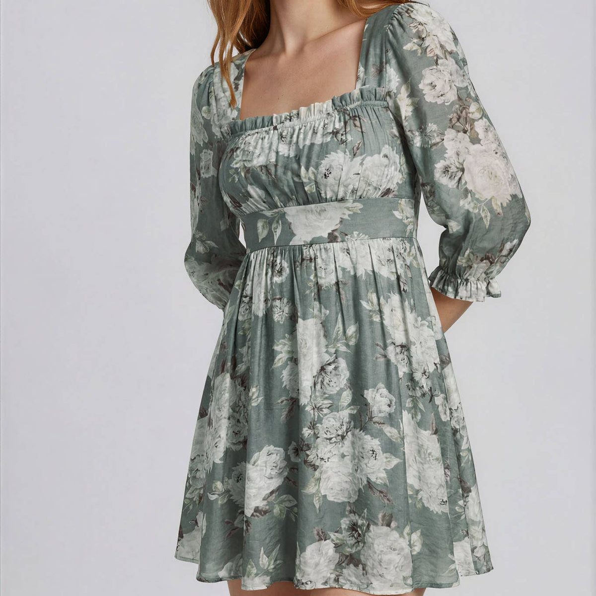 A woman modeling a sage green and white floral print mini dress with a square neckline, smocked bodice, and puffed elbow-length sleeves against a light gray studio background.