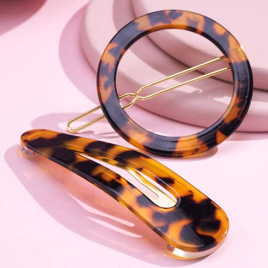 Two tortoiseshell hair clips, one circular and one curved, with gold-tone metal spring clips, displayed on a pink background with stacked pastel props.