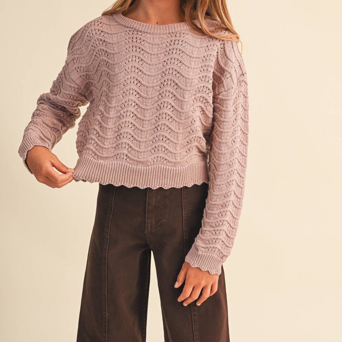 A young girl wearing a dusty pink cable-knit cropped sweater and dark brown wide-leg pants, standing against a solid beige background.