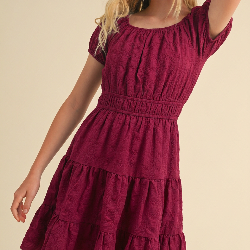 A young blonde woman wearing a short-sleeve tiered dress in deep burgundy, posing against a solid beige background.