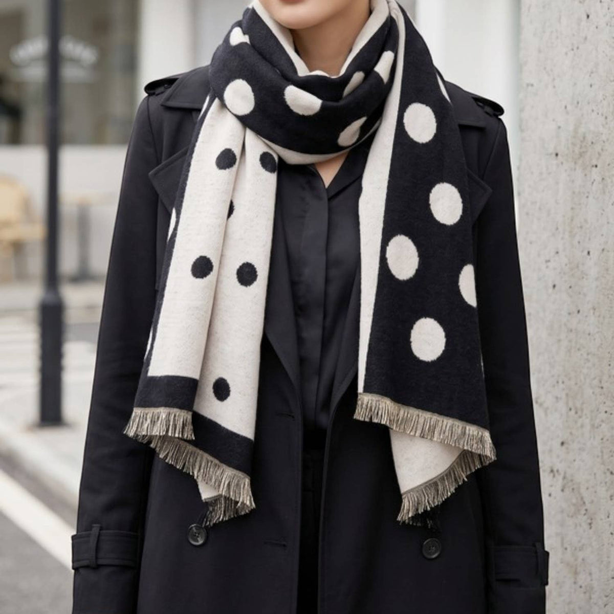 A woman wearing a black and off-white polka dot scarf with fringe, styled over a black coat in an outdoor urban setting.