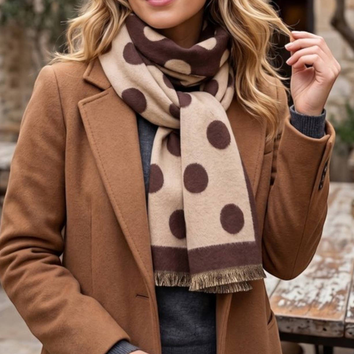 A woman wearing a camel brown coat and a beige scarf with dark brown polka dots, standing in an outdoor stone courtyard setting.