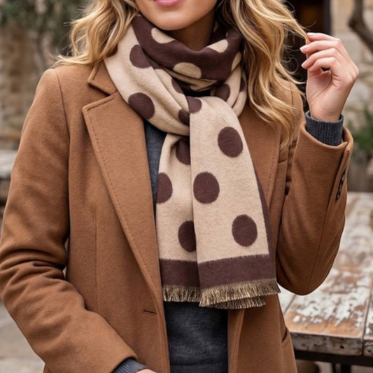 A woman wearing a camel brown coat and a beige scarf with dark brown polka dots, standing in an outdoor stone courtyard setting.