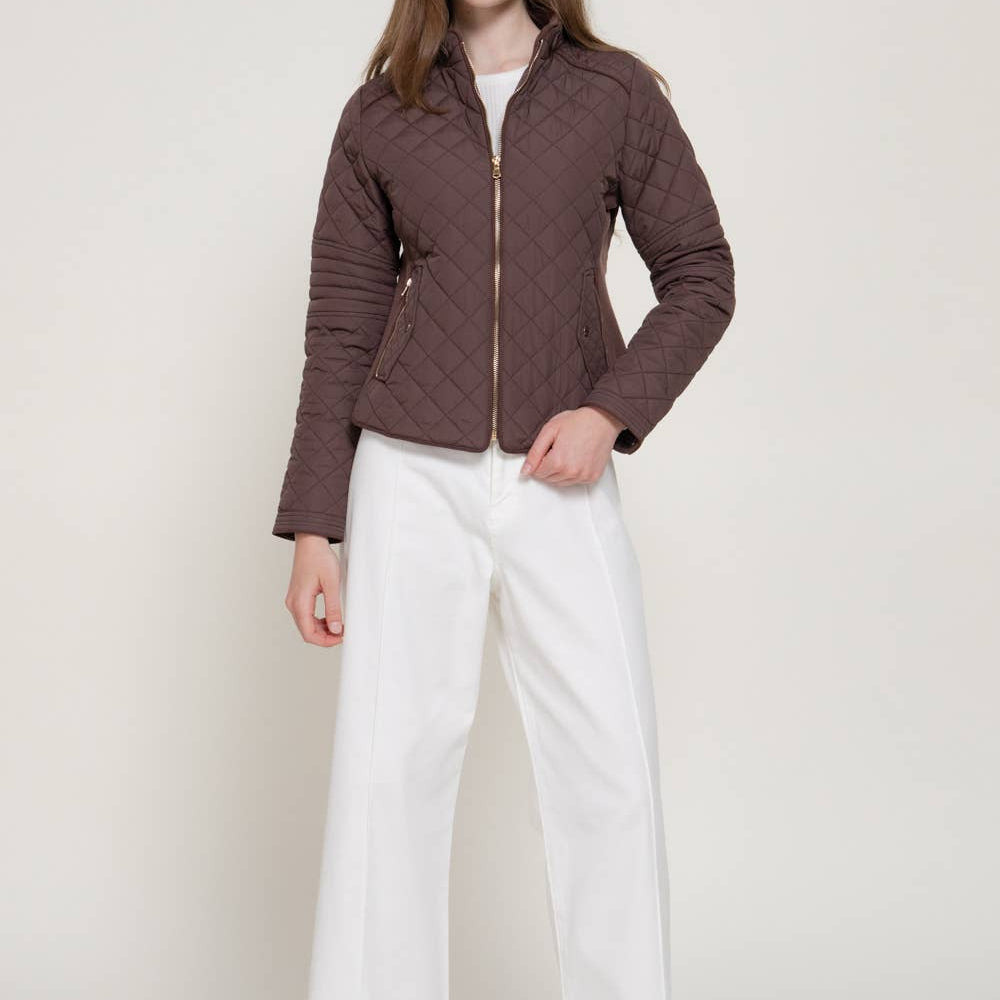 A model wearing a brown quilted zip-up jacket, white wide-leg pants, and black boots, standing against a light beige studio background. A black 'PLUS SIZE' label is in the top right corner.