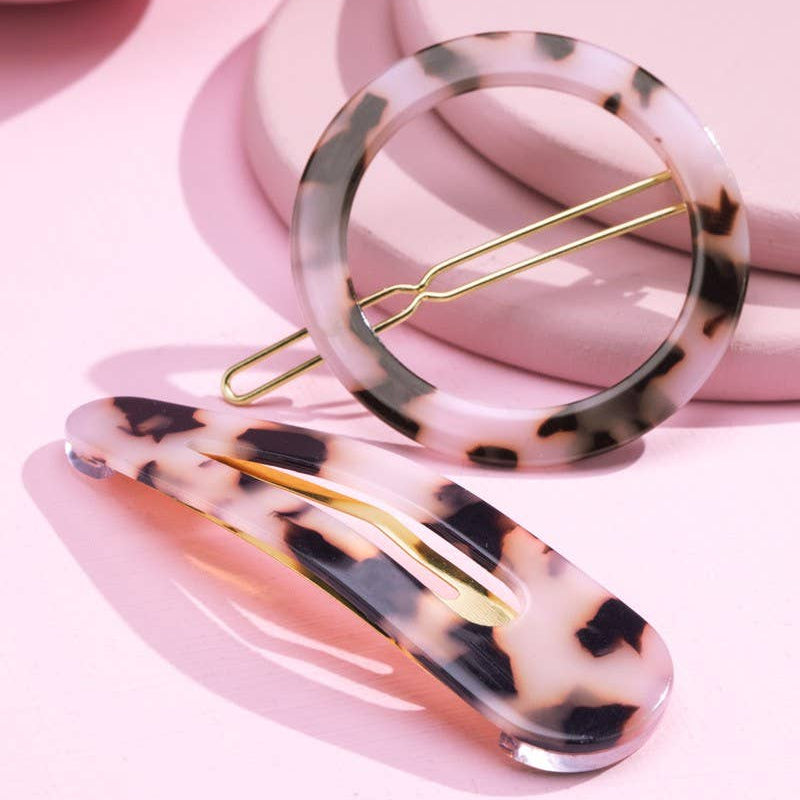 Two tortoiseshell-style hair claw clips with gold-tone metal springs, displayed on a pink background with soft pink geometric props.