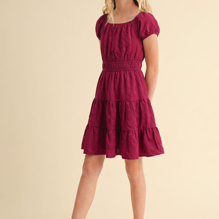 A young girl with blonde hair wearing a tiered burgundy short-sleeve dress and white woven Mary Jane shoes, standing against a solid beige studio background.