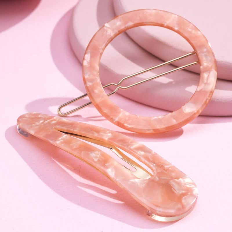 Two peach-pink marbled hair clips with gold metal wire springs, styled on a pink background with soft pastel props.